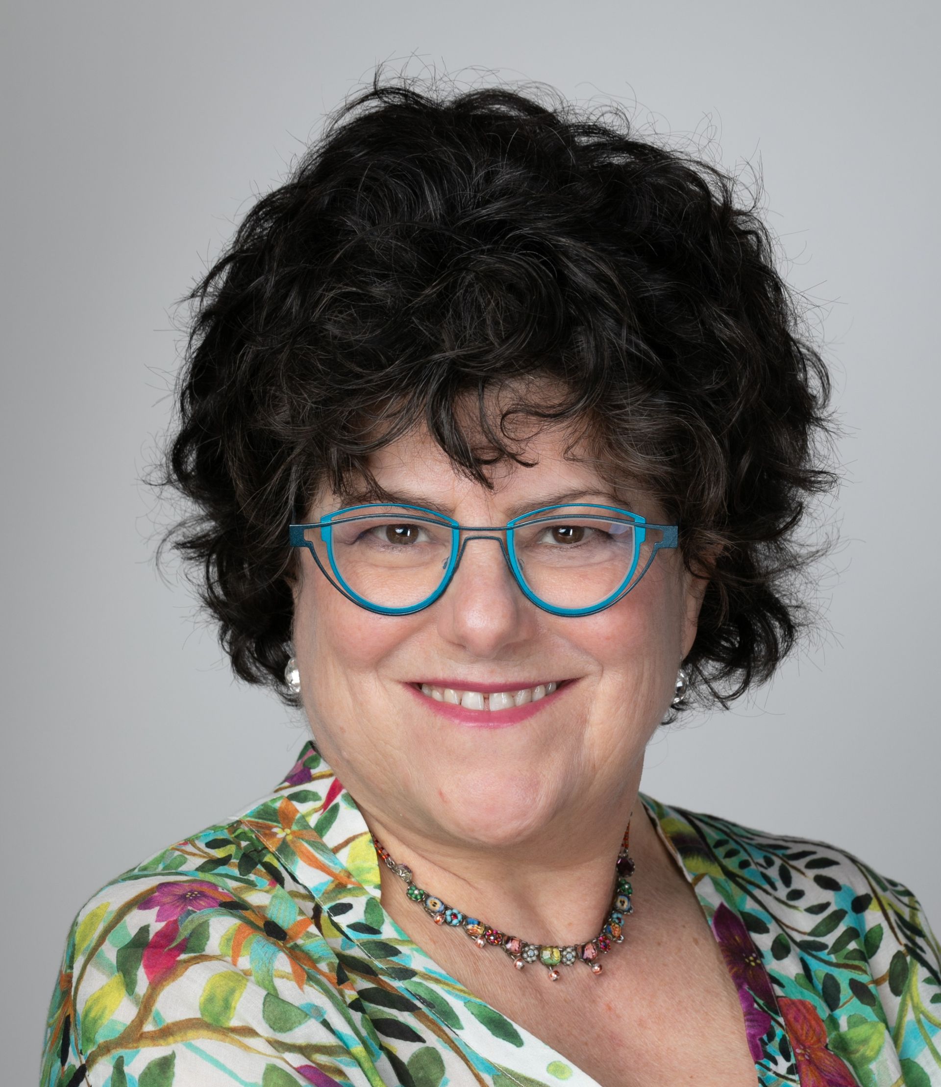 Woman with curly dark hair, turquoise glasses, smiling, wearing colorful floral top.