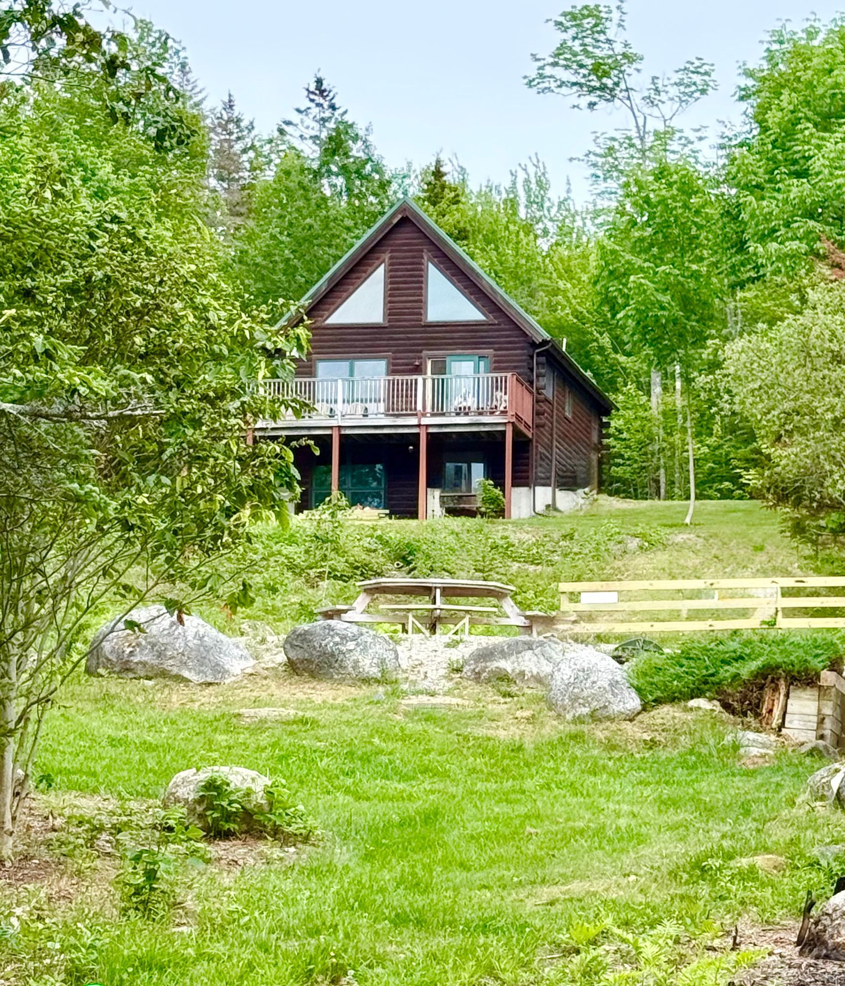 Log cabin with green roof and large deck, surrounded by trees.