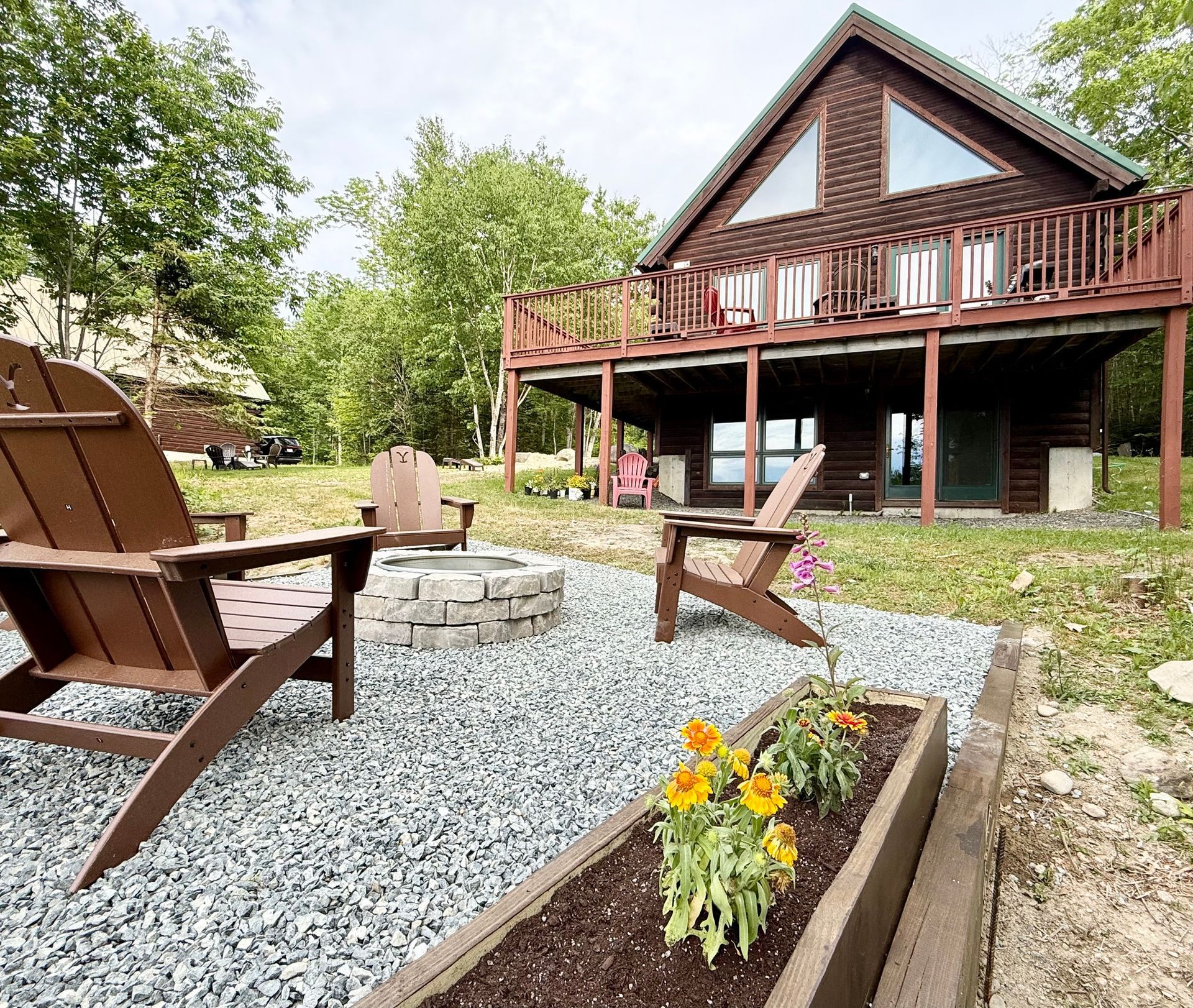 Log cabin with wooden deck, chairs, and surrounding trees in a sunny outdoor setting.