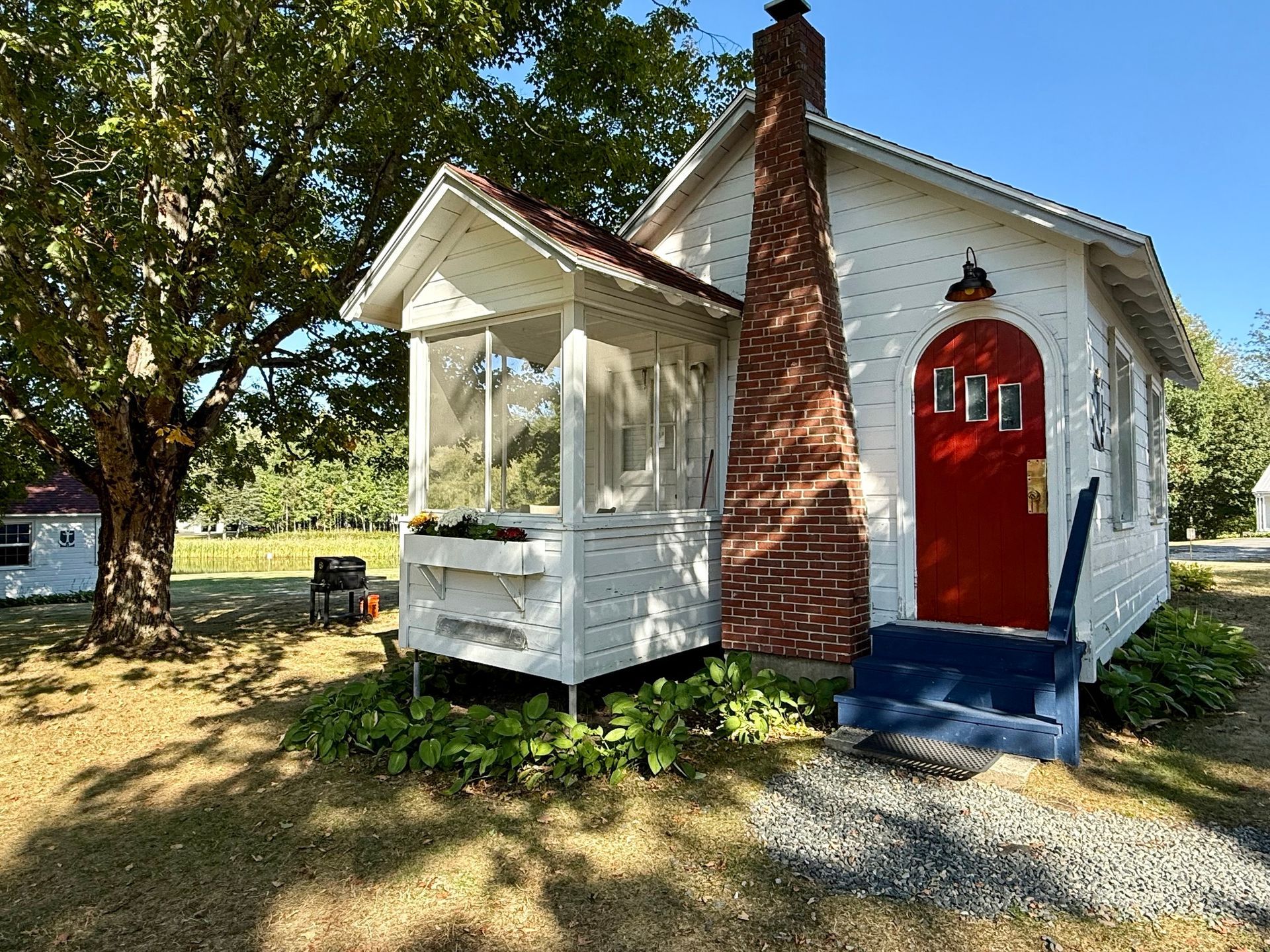 Quaint white house with red door and brick chimney, screened porch, and lush greenery.