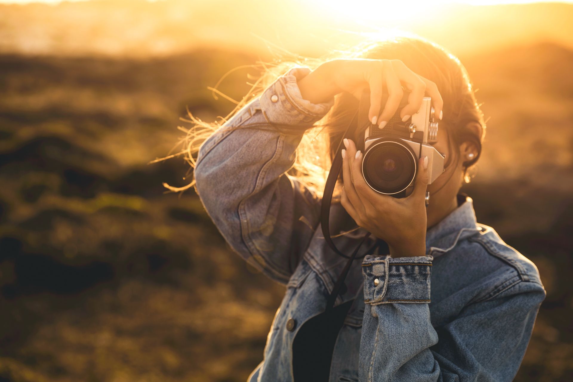 Woman in denim jacket holding camera, taking photo at sunset.