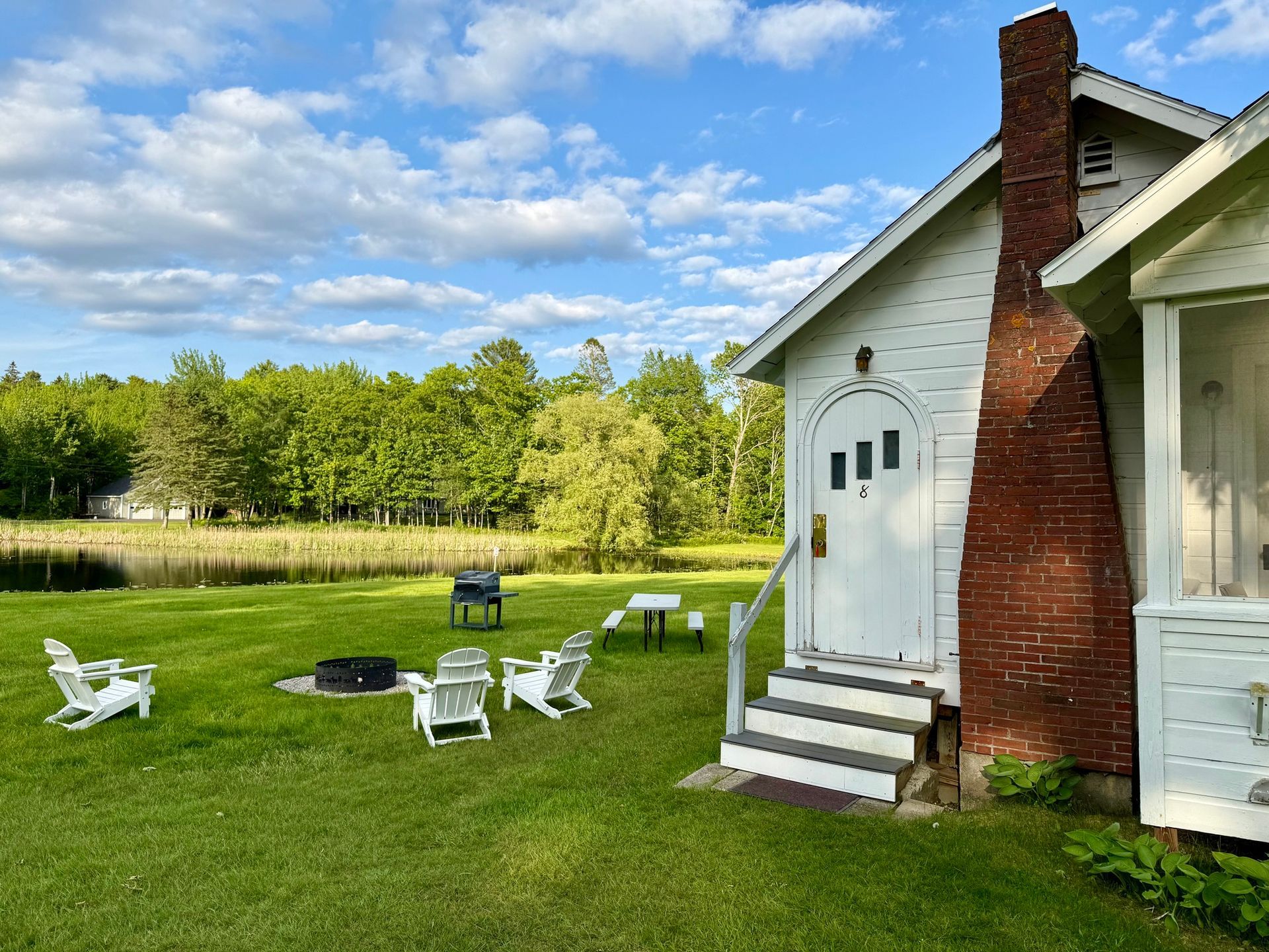 White cottage with a porch sits on a green lawn near trees and a small pond, on a sunny day.