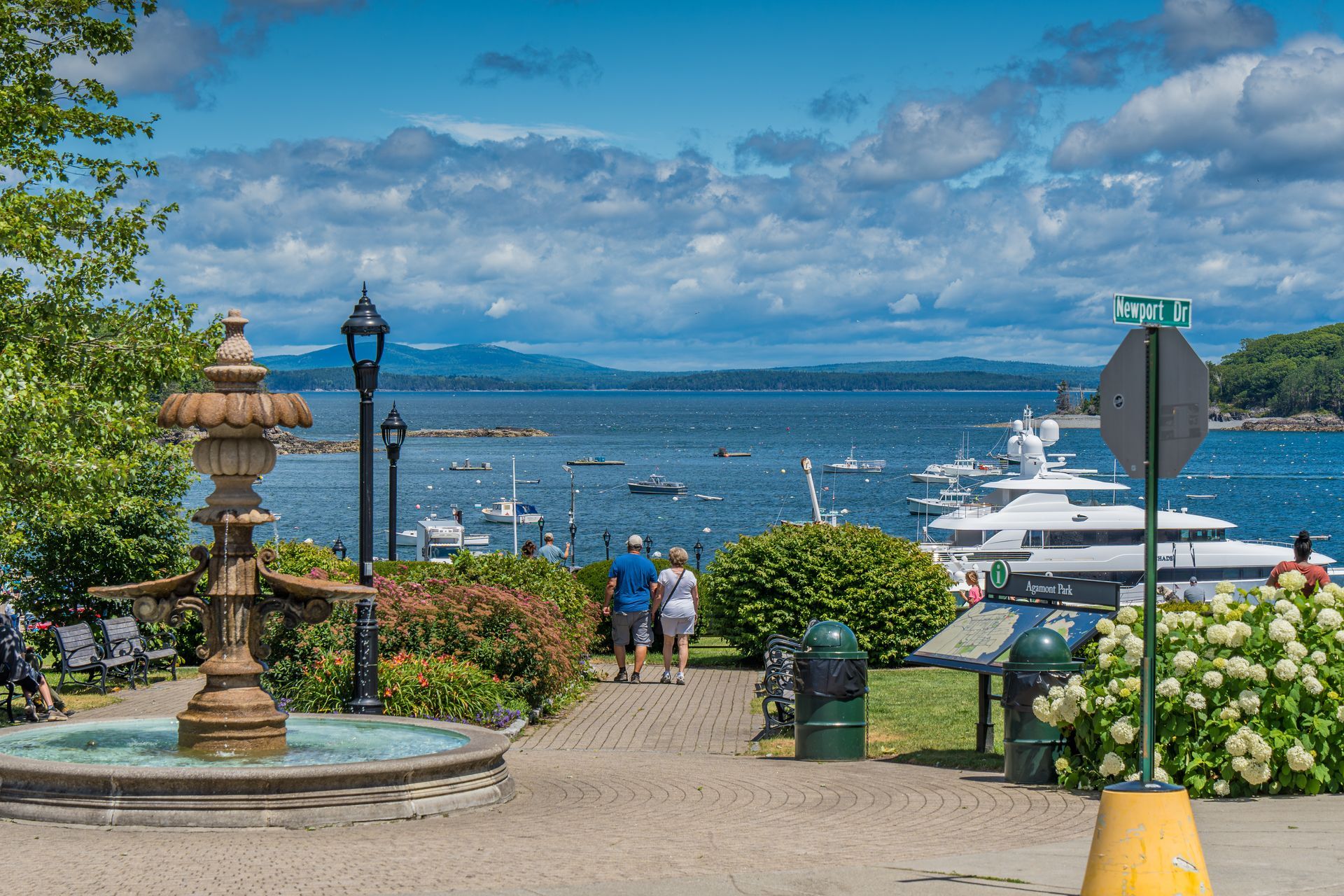 A view of Camden Harbor, Maine. A stone fountain and path lead to the water with boats, mountains, and blue sky.