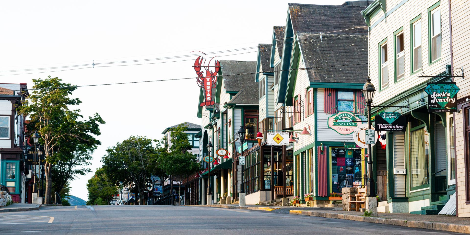 Street lined with shops and restaurants in a coastal town, featuring a large lobster sign, green accents, and a view down the road.