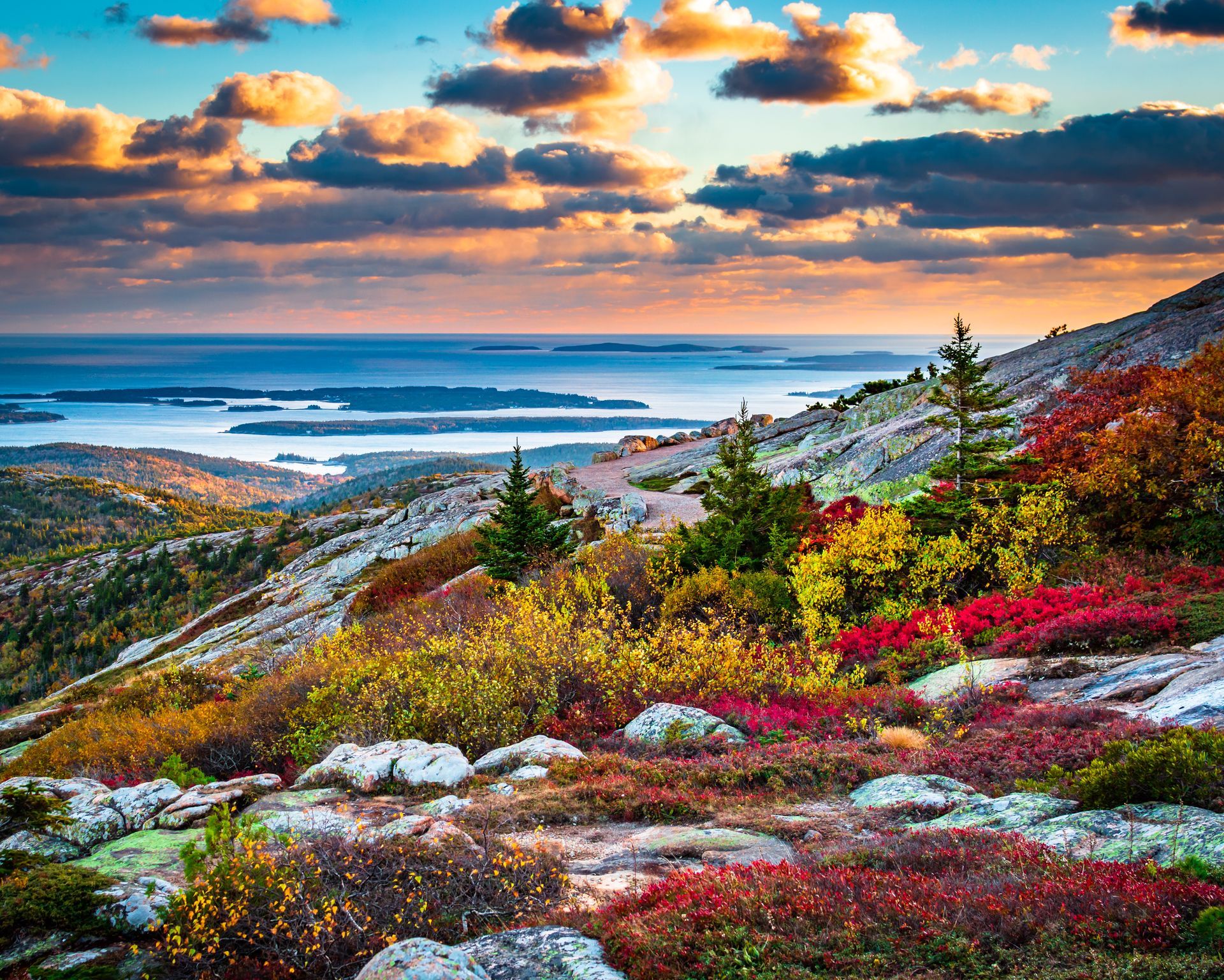 Autumn landscape with colorful foliage on a mountainside overlooking a blue ocean under a sunset sky.