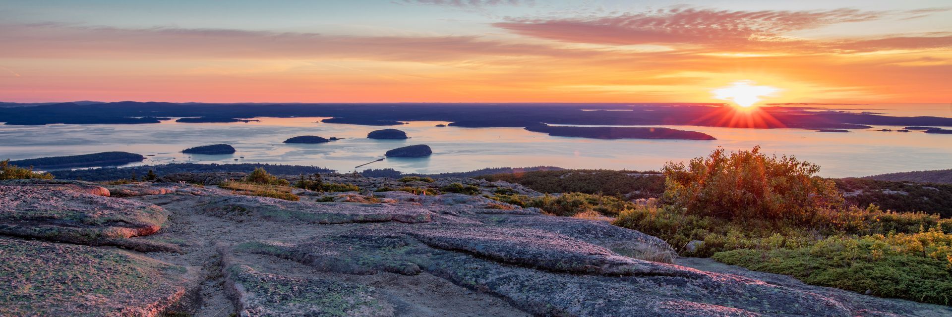 Sunset over ocean and islands from a rocky mountain, with a bright orange sky.