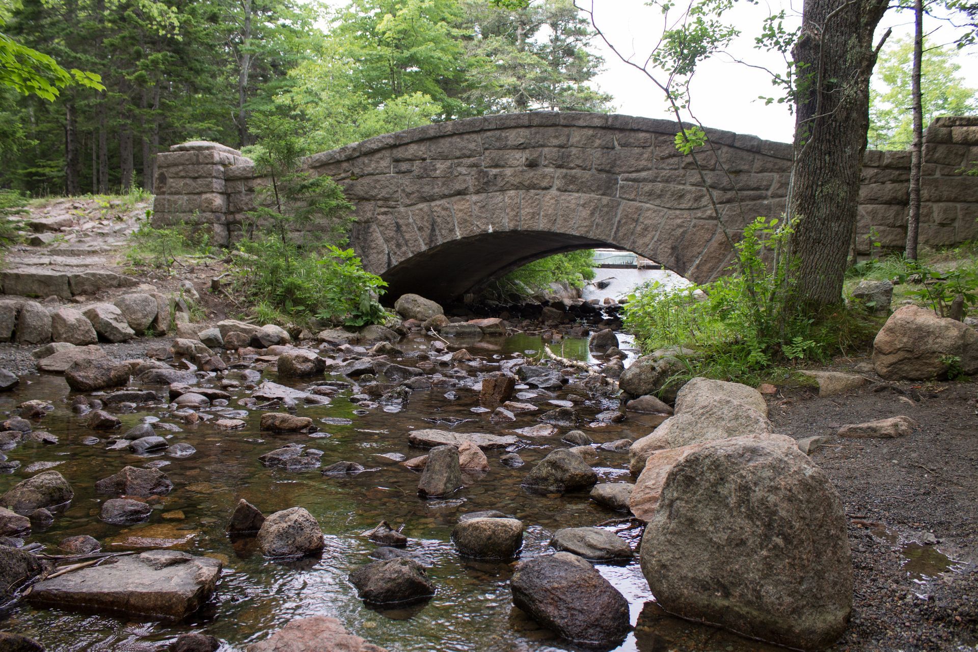 Stone bridge arches over a rocky stream, surrounded by trees.