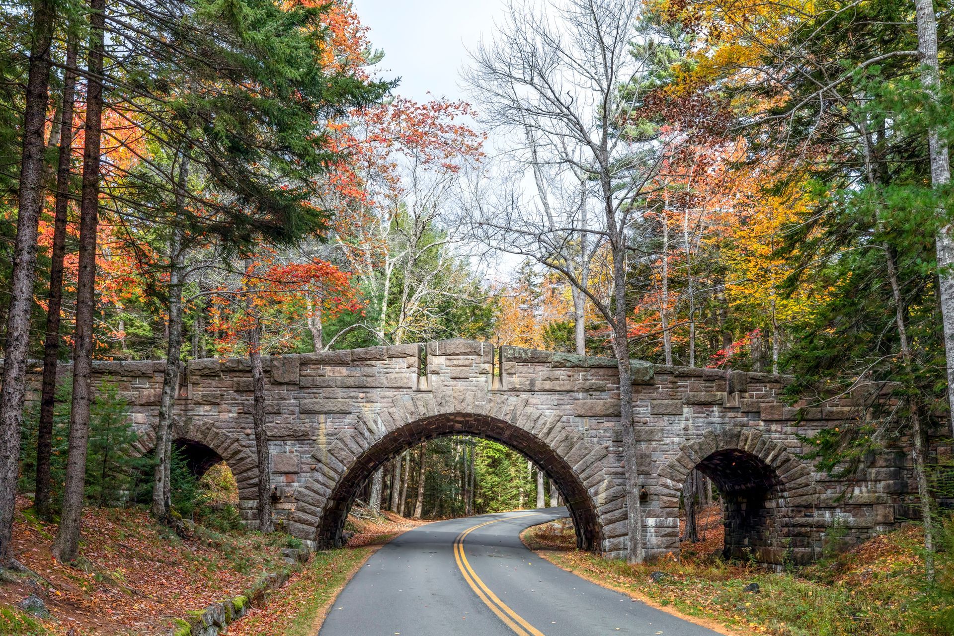 Stone bridge arches over a road through autumn trees with colorful foliage.