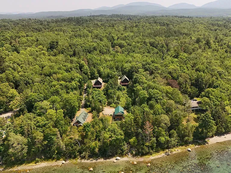 Aerial view of cabins nestled in a dense green forest, bordering a shoreline with clear water.