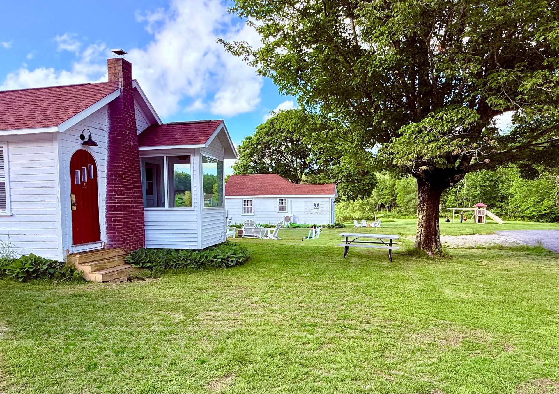 Small white house with a red door beside a large tree on a grassy lawn under a blue sky