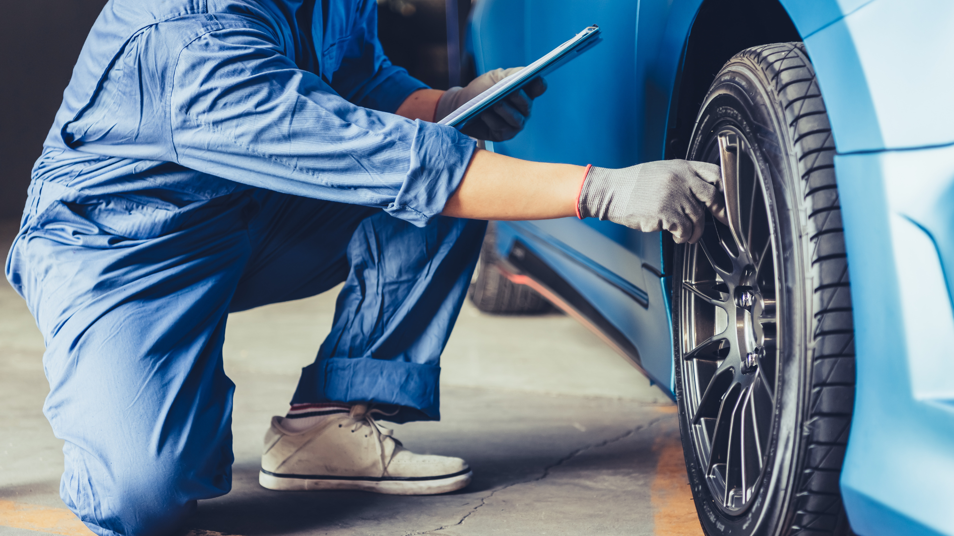 Mechanic in blue overalls inspecting a car tire, holding a clipboard in a garage.