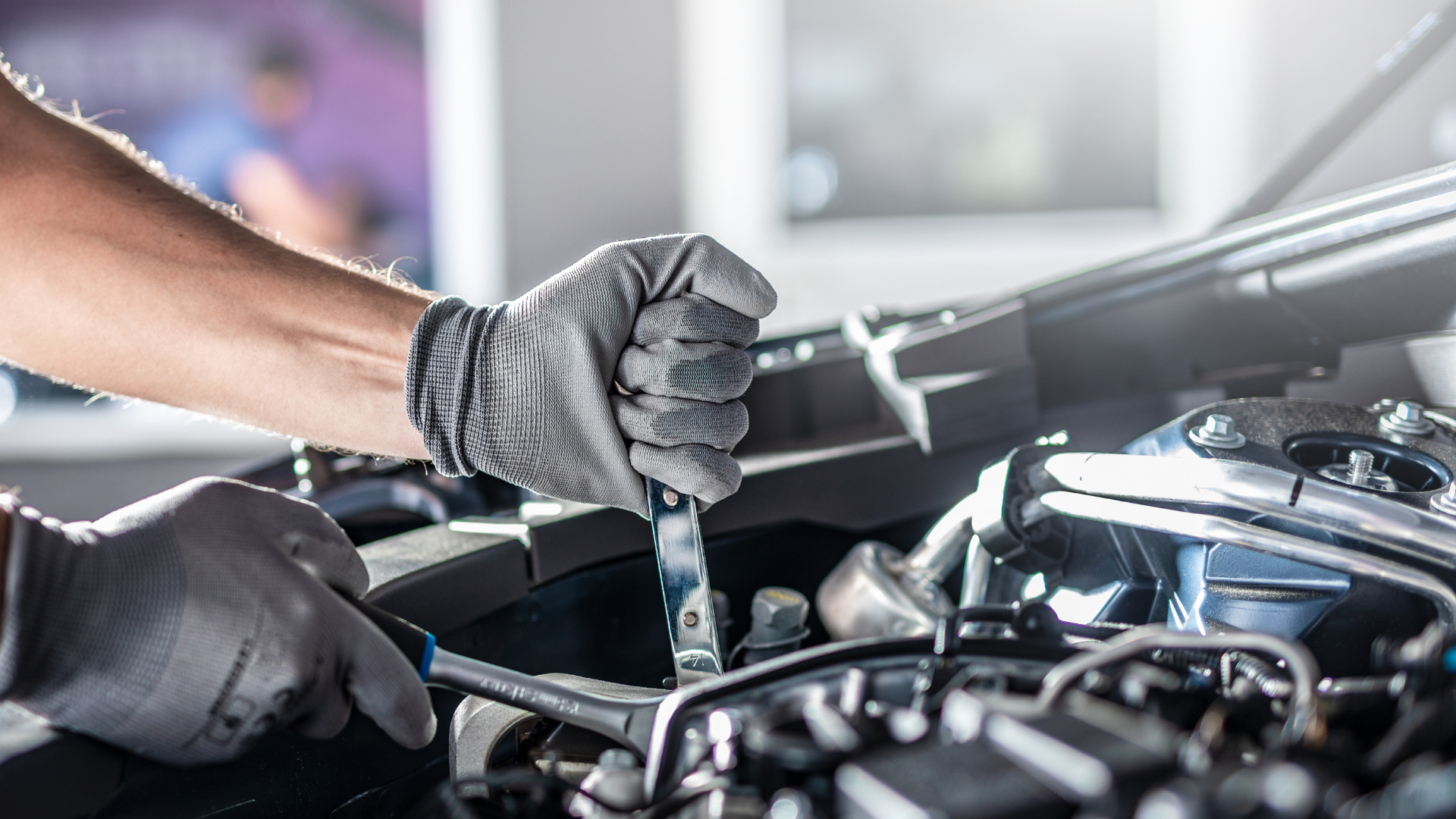 A mechanic in gray gloves works on a car engine with a wrench in a well-lit garage.