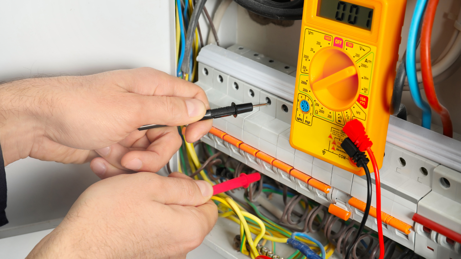 Electrician using a multimeter to test electrical wiring inside a distribution panel; hands, wires, and device are visible.