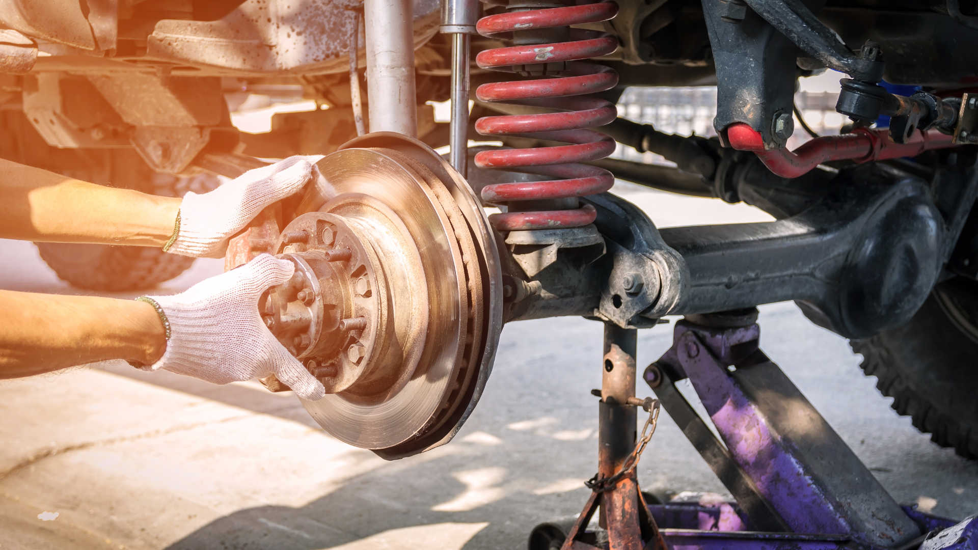 Hands in gloves replacing a car's brake rotor, with the vehicle lifted by a jack.