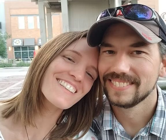 Smiling couple, woman with long brown hair, man in a cap and mustache, close together outdoors.