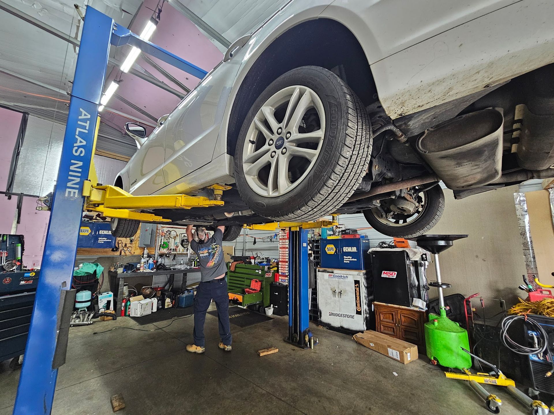 Mechanic working under a white car lifted on a blue and yellow car lift inside a cluttered garage.