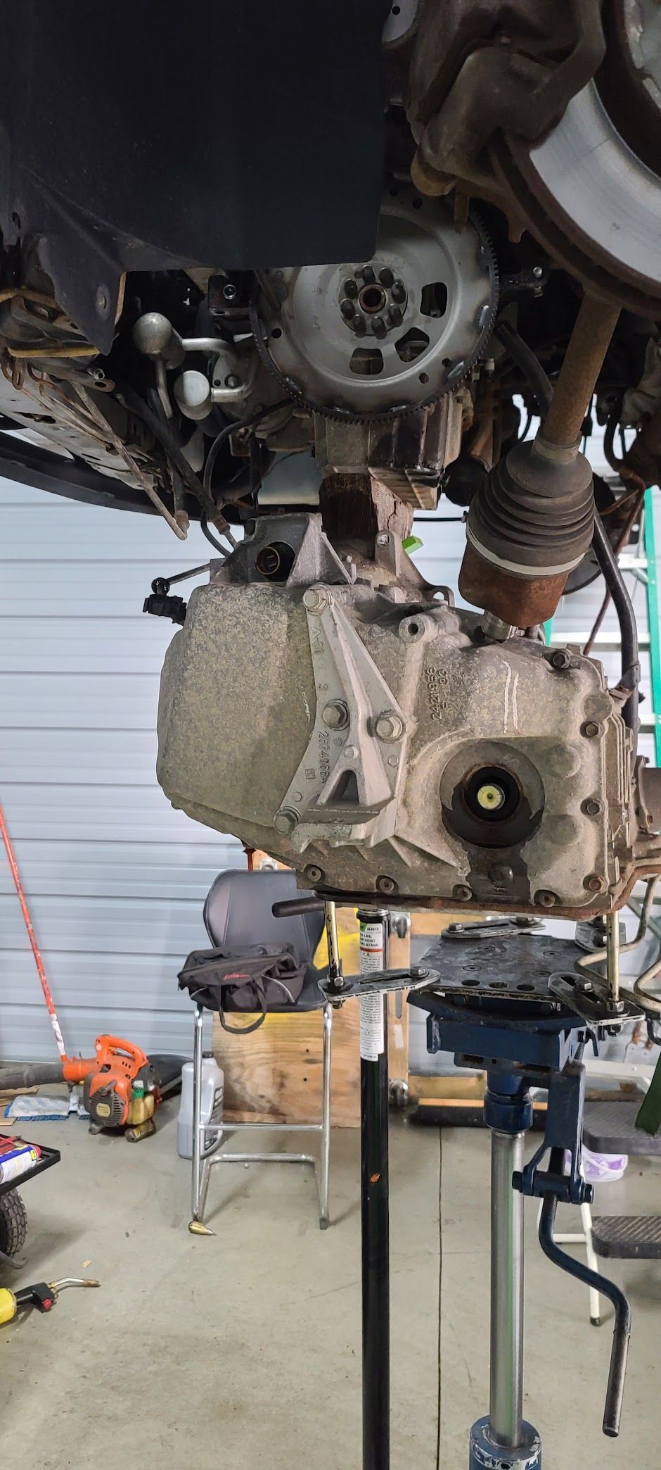 A car transmission being worked on, supported by a jack. The car is in a garage.
