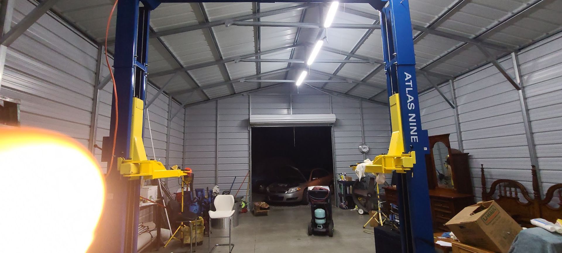An auto shop with a car lift in the center. Open garage door in the background, tools, and furniture around the perimeter.