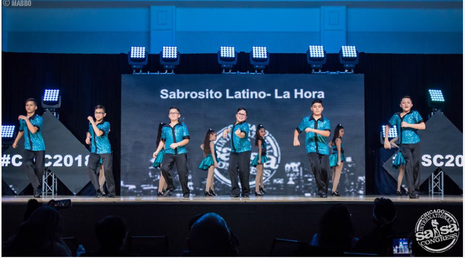 A group of people are dancing on a stage in front of a large screen.