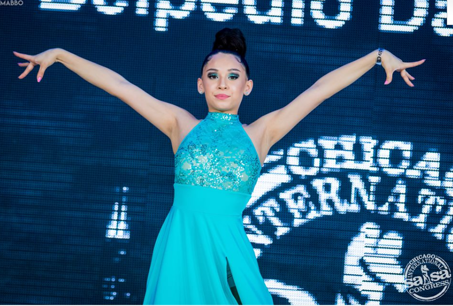 A woman in a blue dress stands in front of a screen that says chica terna