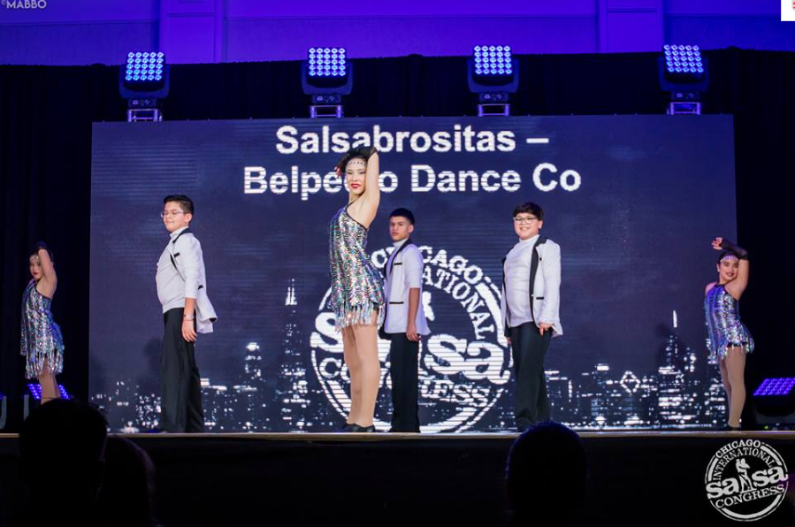 A group of children are performing on a stage in front of a large screen that says salsabrositas
