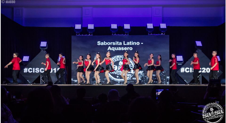 A group of people are dancing on a stage in front of a large screen.