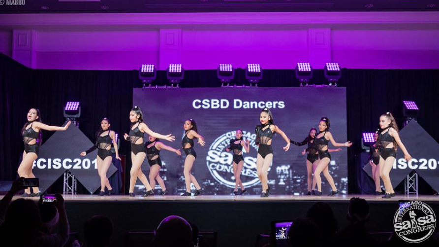 A group of dancers are performing on a stage in front of a large screen.