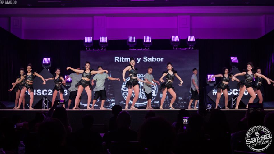 A group of people are dancing on a stage in front of a large screen.