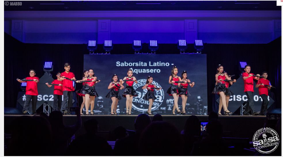 A group of people are dancing on a stage in front of a large screen.