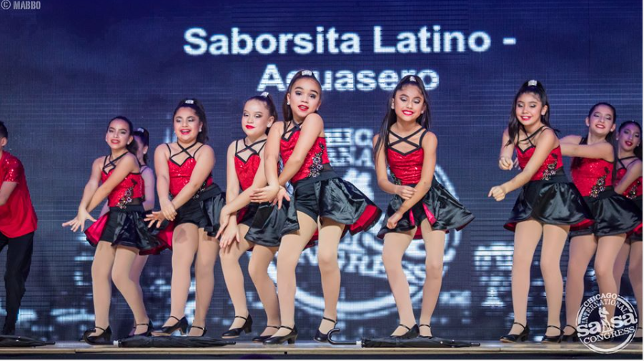 A group of young girls are dancing in front of a sign that says saborsita latino