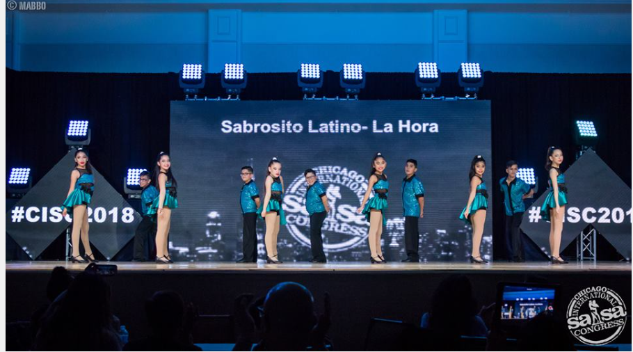A group of people are dancing on a stage in front of a large screen.