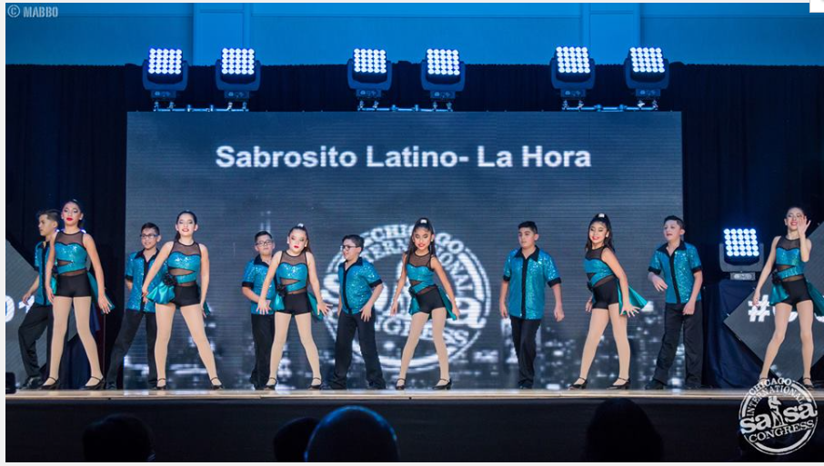 A group of kids are dancing on a stage with a sign that says sabroso latino-la hora