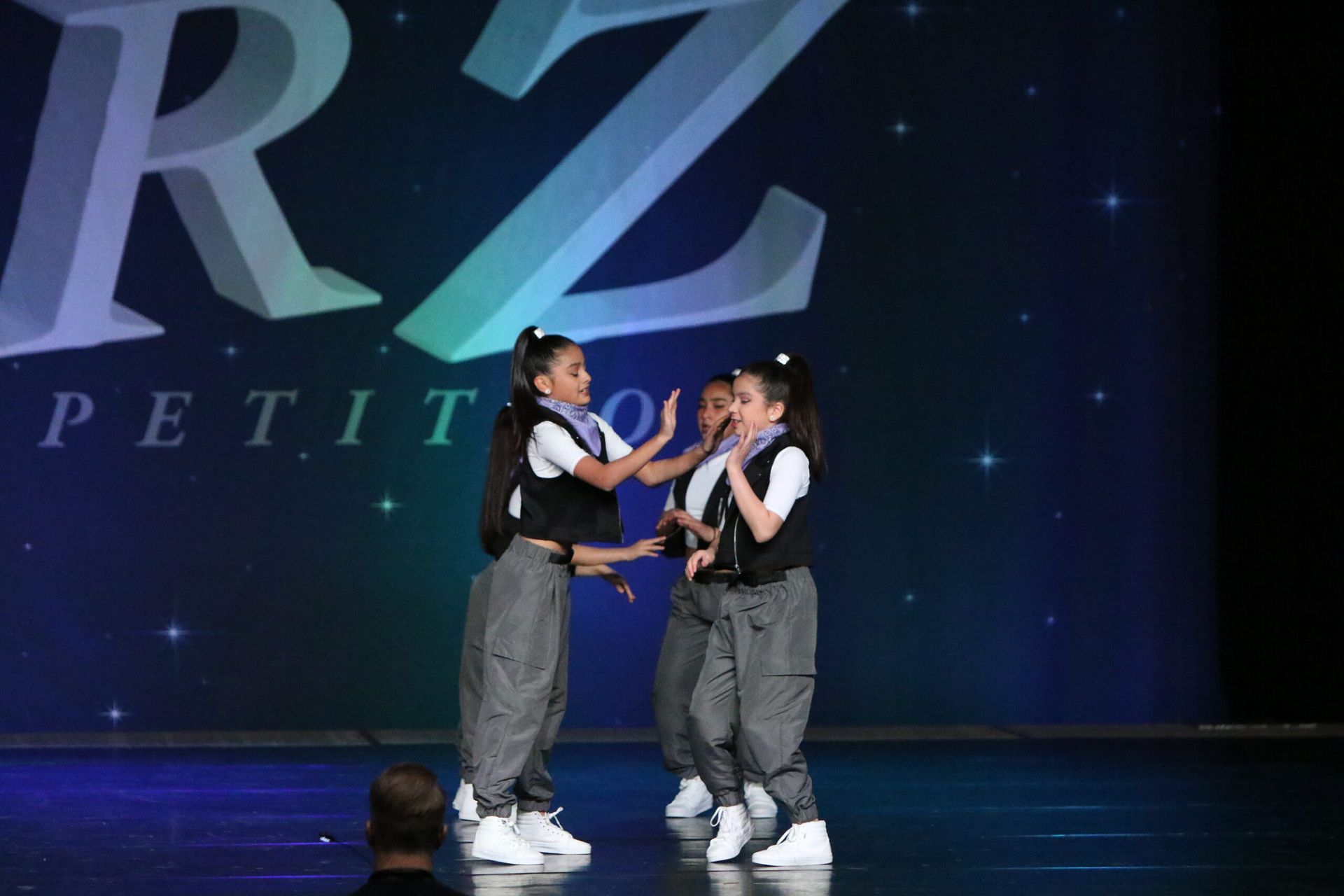 Three young girls are dancing on a stage in front of a rz logo.
