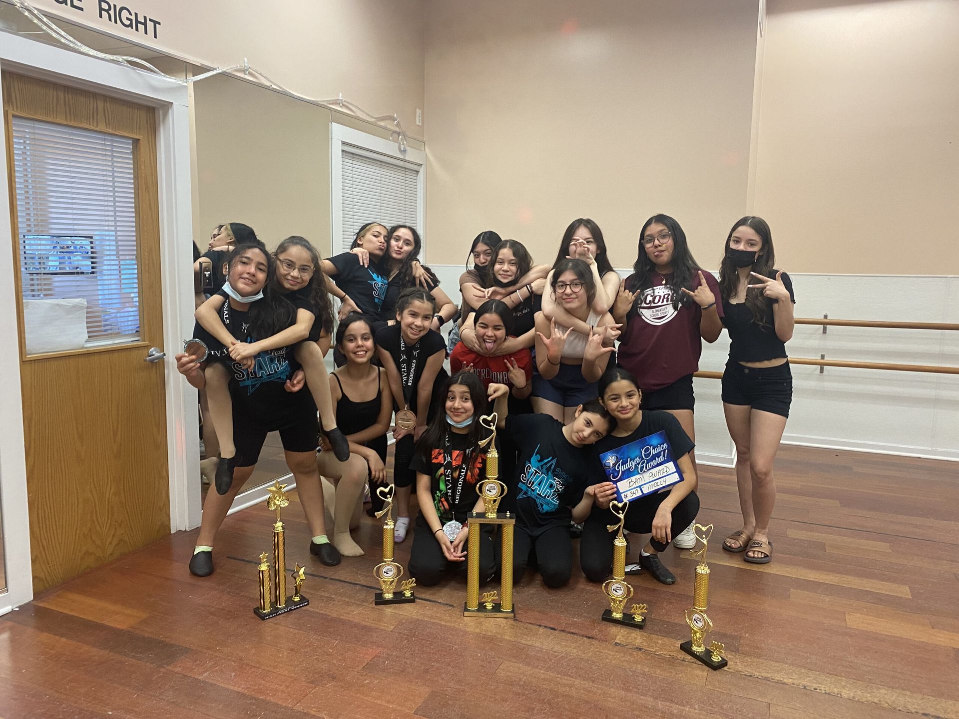 A group of young people are posing for a picture in a dance studio with trophies.