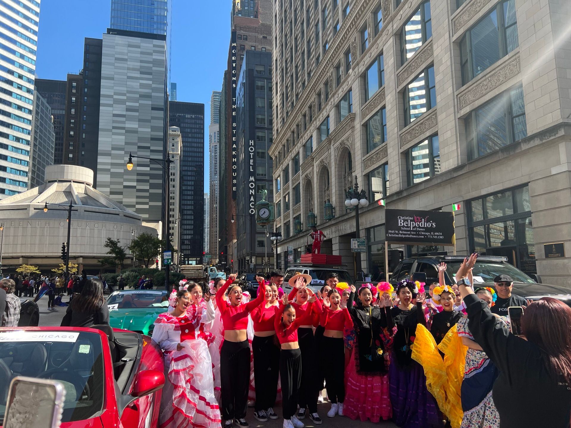 A group of people are standing in front of a red car on a city street.