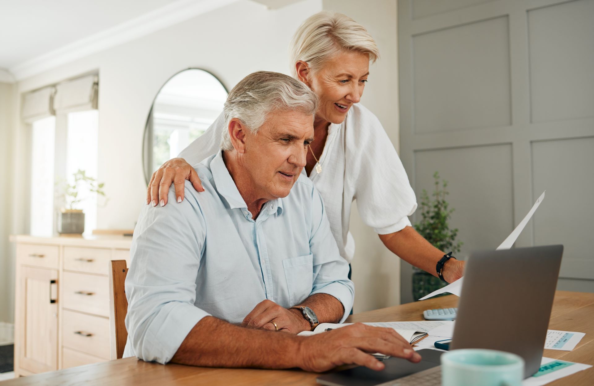 Couple reviewing financial documents on a laptop at a table. Woman has arm on man's shoulder.