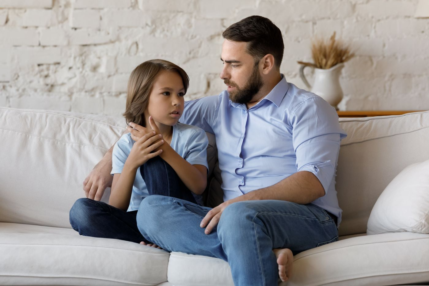 Man with beard comforts child on a white couch; both are looking at each other, one hand on the child's shoulder.