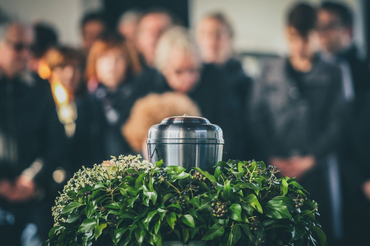 Urn on a green wreath at a memorial service, people in the background bowing heads in mourning.