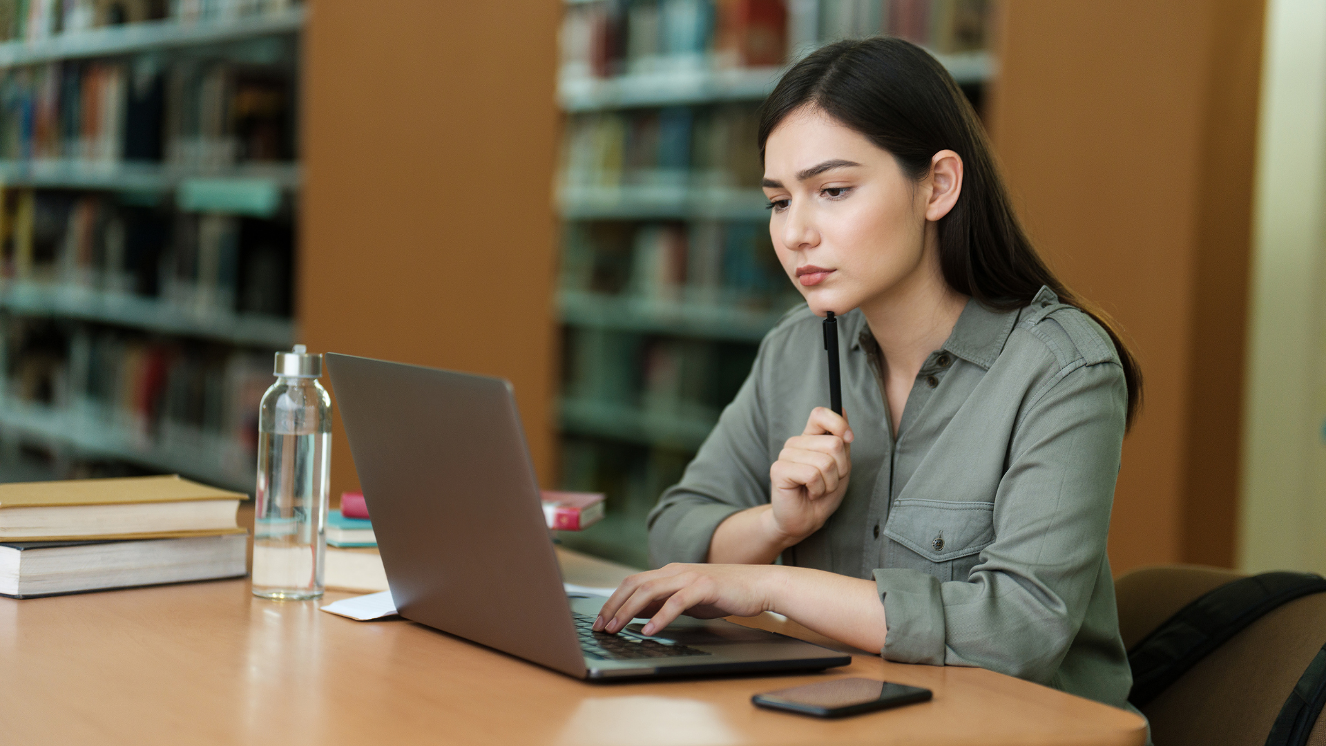 Woman working on a laptop at a library table with books and a smartphone nearby.