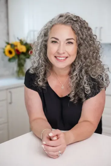 Smiling woman with curly gray hair leaning on a white table in a bright kitchen
