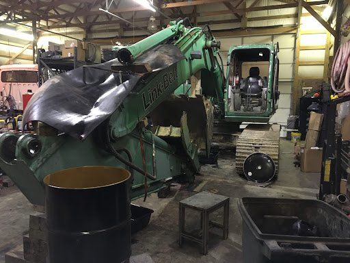 A green excavator is sitting in a garage next to a black barrel.