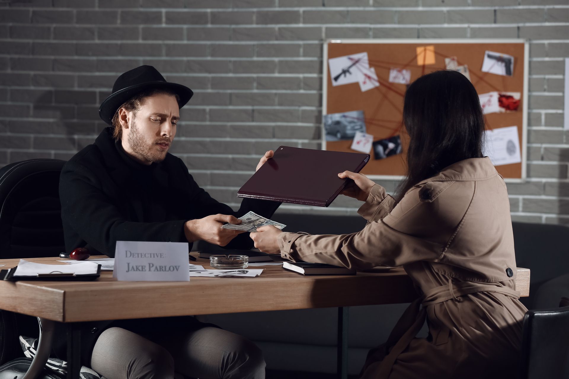 A man in a hat receives cash from a woman across a desk in a detective's office.