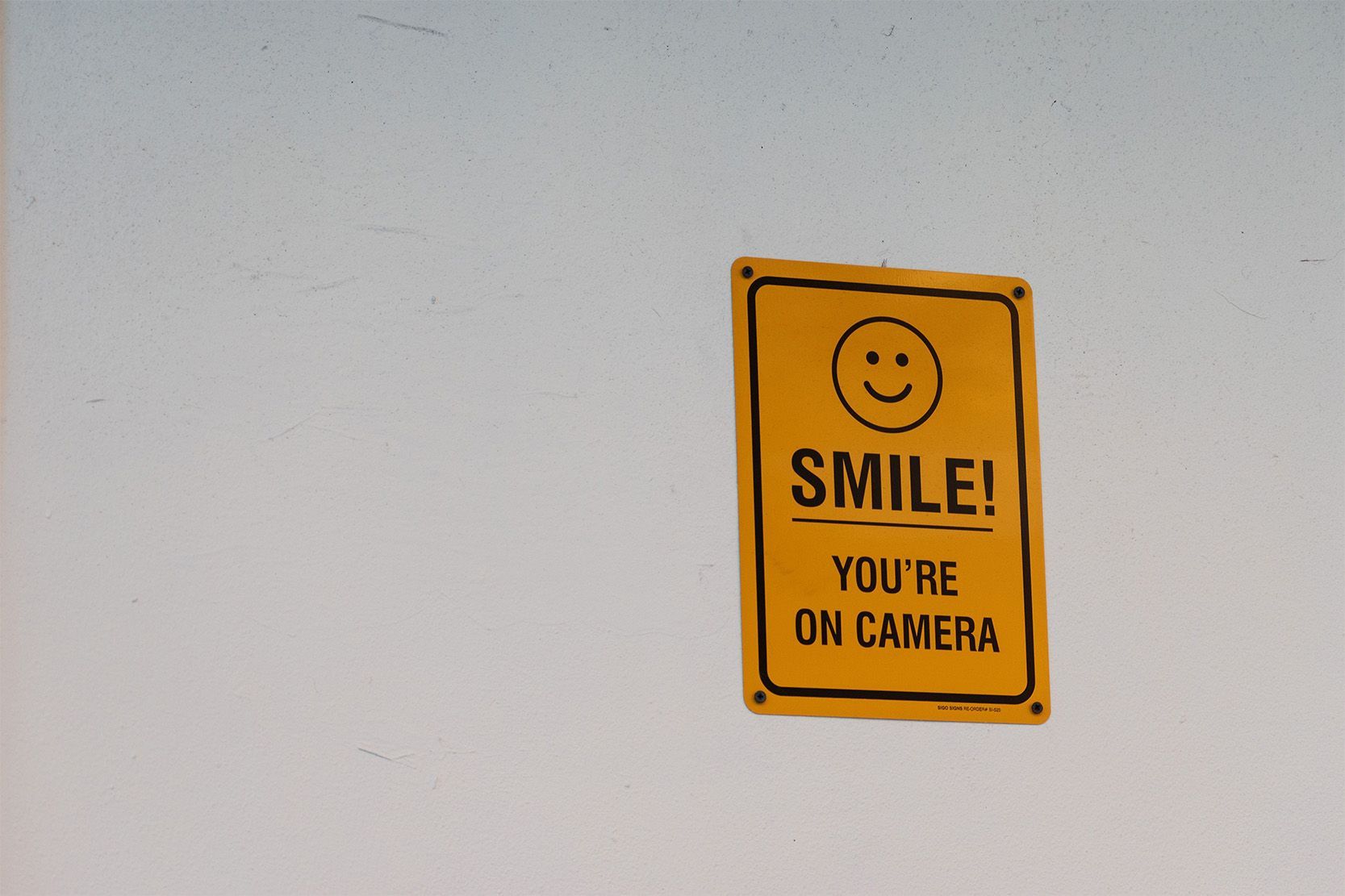 Yellow “Smile! You’re on camera” video surveillance sign mounted on a white wall.