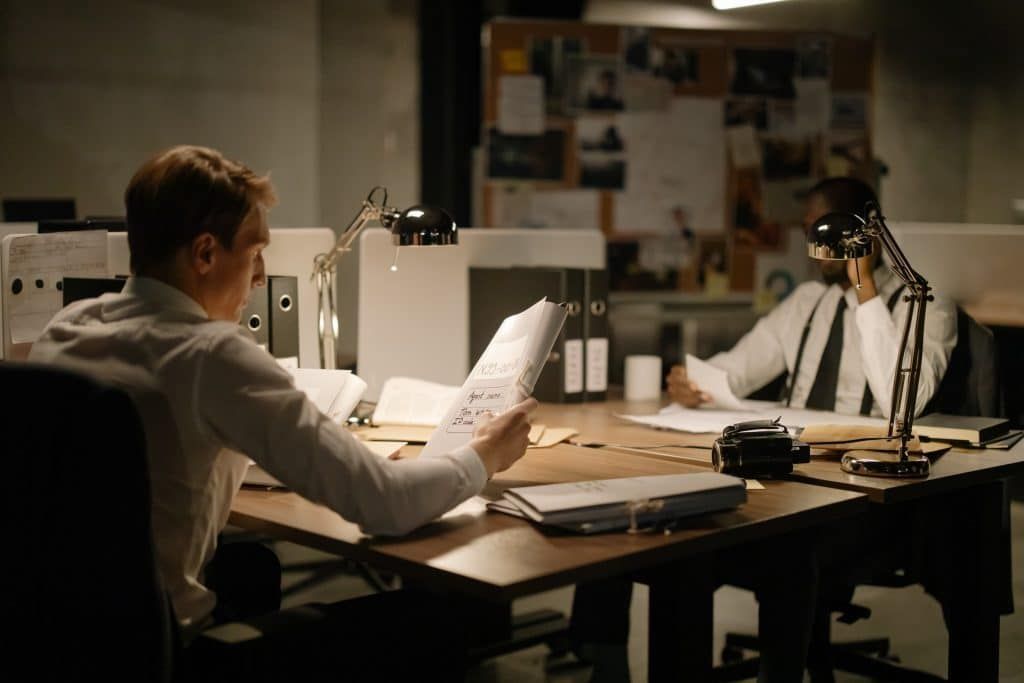 Two private investigators analyzing case files at a desk covered with documents, photographs, and investigation notes.
