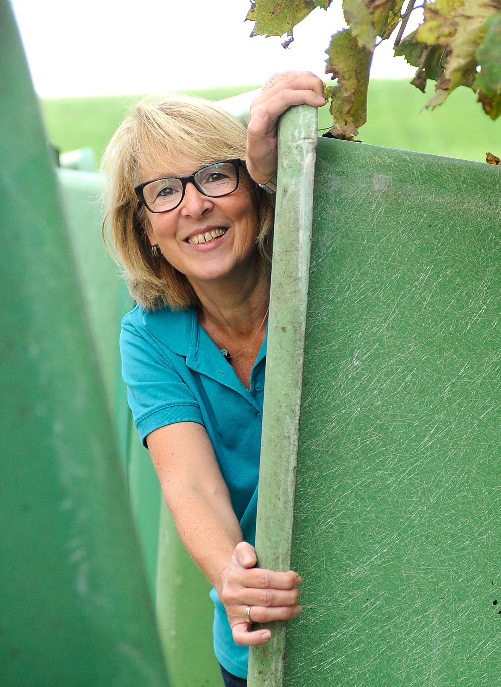 Eine Frau mit Brille späht hinter einem grünen Zaun hervor.