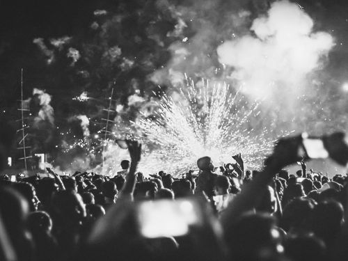 Foule à un concert avec feux d'artifice. Les gens lèvent les bras, tenant leurs téléphones dans le noir. Photo en noir et blanc.
