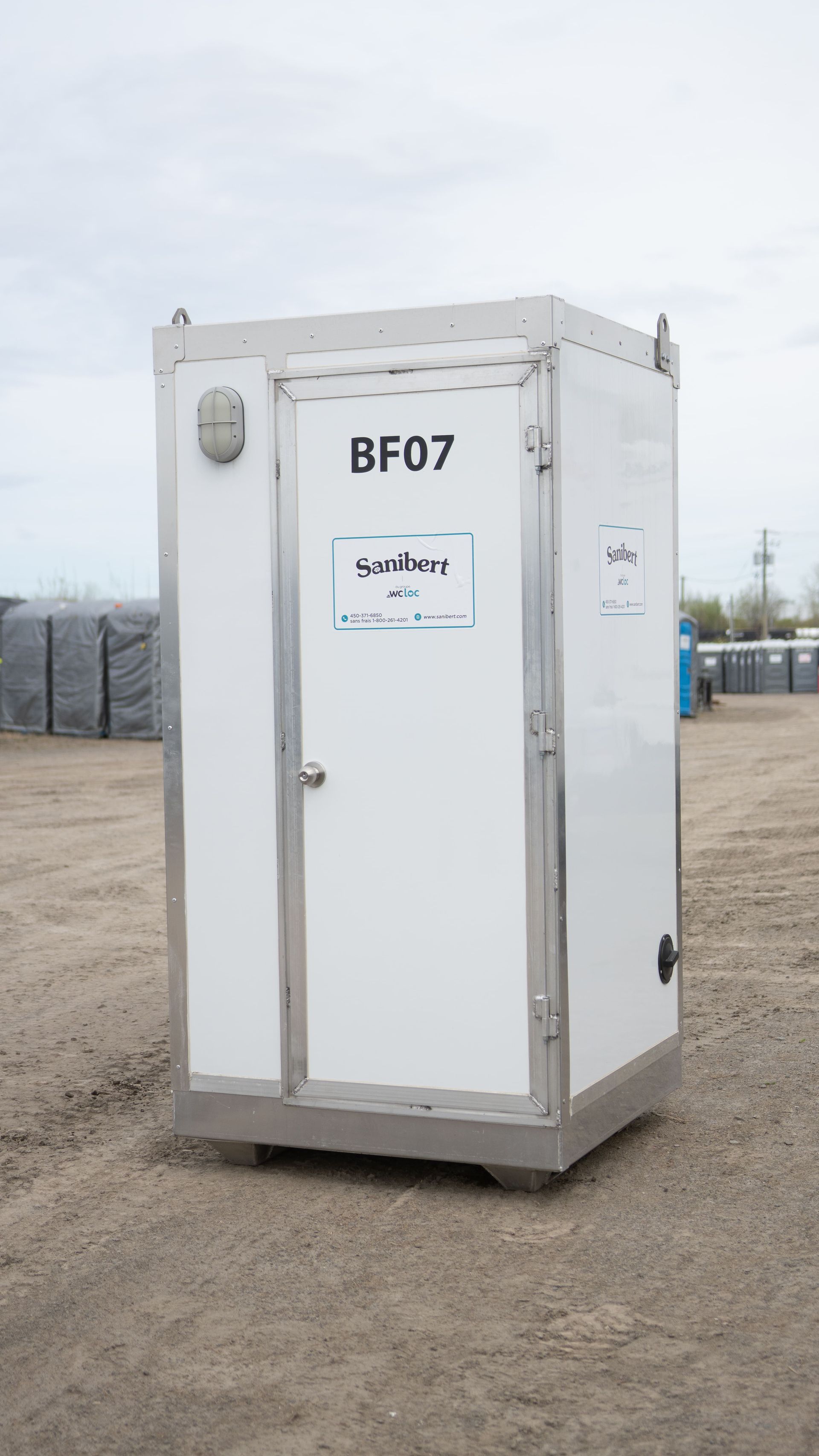 Toilettes portables blanches avec l'inscription « BF07 » sur la porte, à l'extérieur sur du gravier, avec d'autres toilettes en arrière-plan.