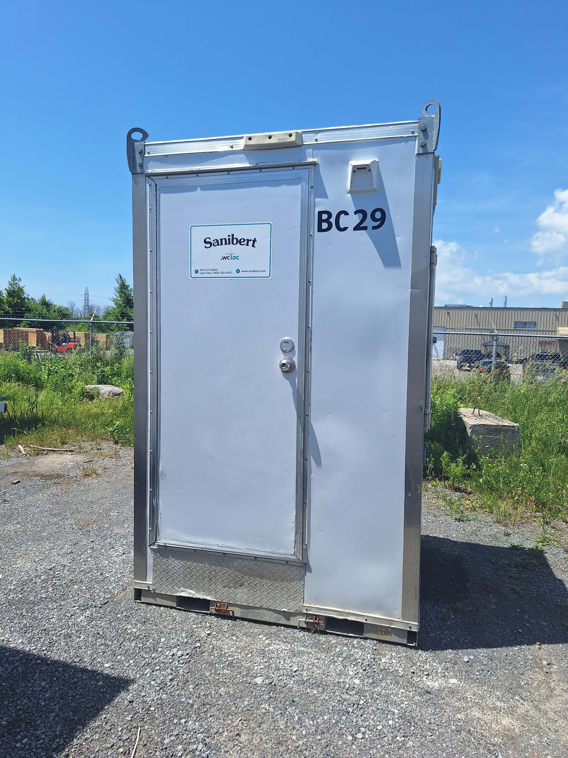 Toilettes portatives argentées dans un terrain en gravier par une journée ensoleillée; « BC29 » sur le côté.