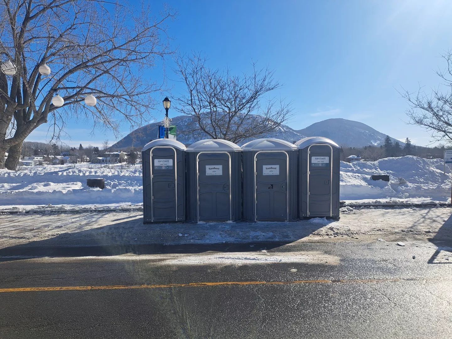 Rangée de toilettes portables grises dans un décor extérieur enneigé ; montagnes et arbres en arrière-plan par une journée ensoleillée.