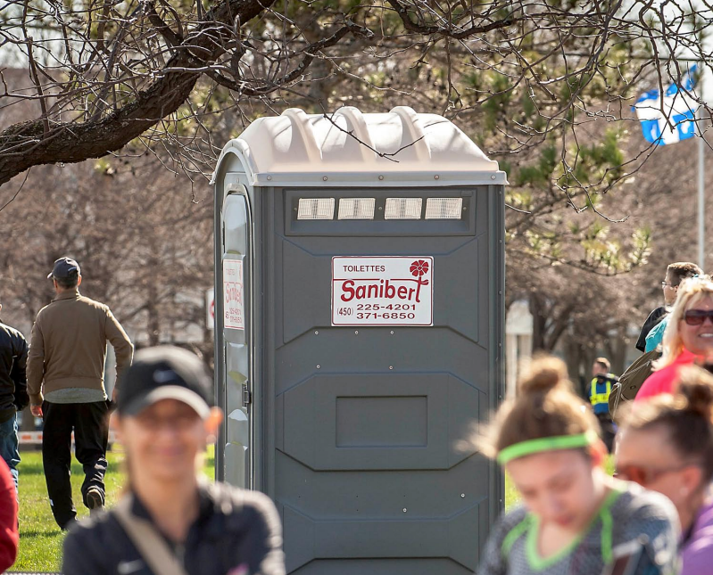 Toilettes portables grises avec logo rouge dans le parc, personnes marchant et souriant.